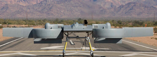 A grey drone with twin engines sits on a runway in a desert landscape with mountains in the background.