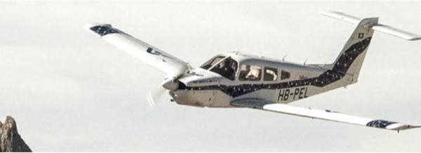 A small white aeroplane with blue accents and the registration HB-PEL flies in the sky near a mountain peak.