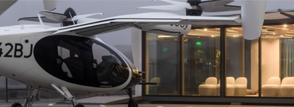 A white electric vertical take-off aircraft is parked outside a modern glass building with white chairs inside.